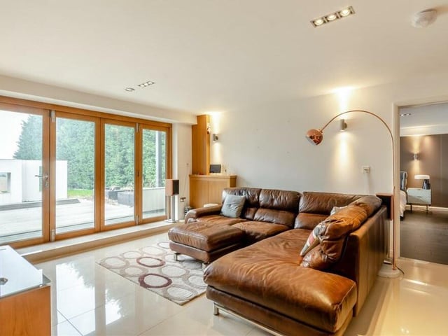 As we move downstairs to the lower ground floor, the main reception space is this living room, with its large, glazed doors that lead out to the decked patio in the back garden. It boasts a feature wall, porcelain tiled floor and a custom oak cabinet, housing the underfloor heating manifold.