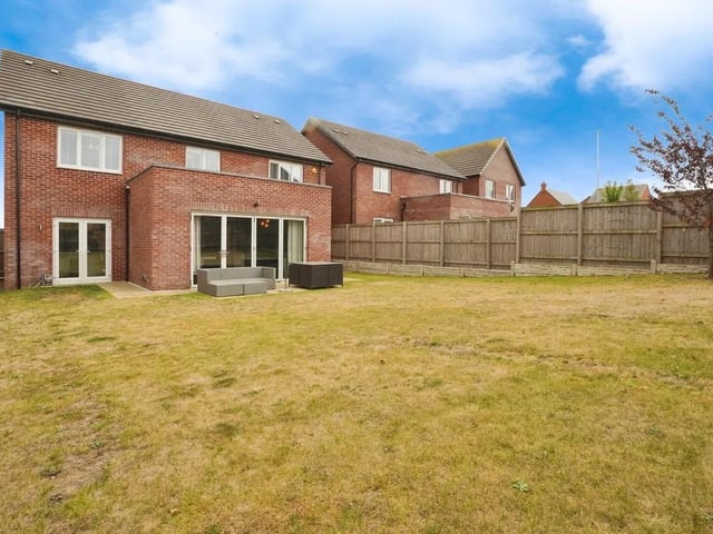 The rear garden offers a generous lawn enclosed by wooden fencing, providing a private and secure outdoor space. There is a paved patio area adjacent to the house, ideal for seating and outdoor entertaining, while the enclosed garden is mainly laid to grass with some shrubbery and trees visible beyond the fence.