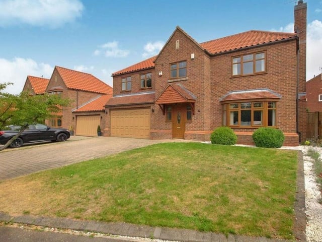 Time now to take a look outside, and this image shows the front of the house. A large, block-paved driveway leads to a double garage and part-borders a generously-sized front lawn, with shrubs and plants.