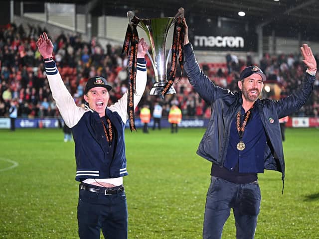 US actor and Wrexham owner Rob McElhenney (L) and US actor and Wrexham owner Ryan Reynolds (R) celebrate on the pitch with the National League trophy after the English National League football match between Wrexham and Boreham Wood at the Racecourse Ground Stadium in Wrexham, north Wales, on April 22, 2023. - Hollywood stars Ryan Reynolds and Rob McElhenney embraced in celebration as they watched Wrexham, who the duo bought in 2020, reach the English Football League for the first time in 15 years on Saturday after a 3-1 win over Boreham Wood. (Photo by Oli SCARFF / AFP) (Photo by OLI SCARFF/AFP via Getty Images)