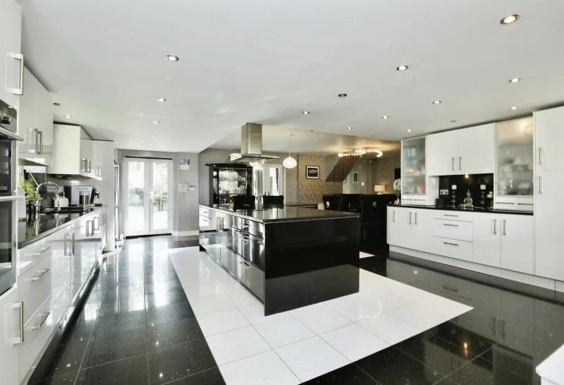 Here is another sparkling, high-gloss, open-plan kitchen. Black granite work surfaces complement a range of modern, white wall and base units. There is an integrated double oven and microwave, five-ring induction hob, dishwasher, washing machine, wine fridge, inset spotlights and granite floor tiles. A central island has cupboards beneath.