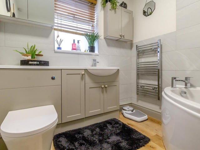 The main bathroom comprises a modern three-piece white suite with chrome fittings. It features a vanity unit with inset wash hand basin and waterfall mixer tap, low-flush WC, work surfaces, storage cupboards, partly tiled walls and luxury vinyl floor.