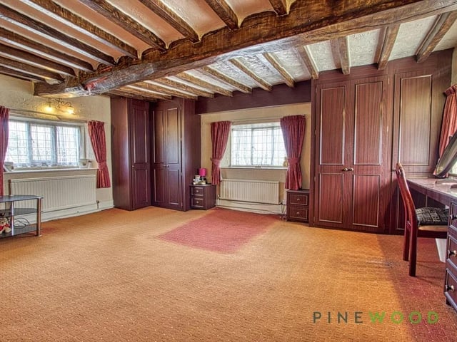 The first-floor master bedroom is a generous and comfortable space featuring exposed beams and stone walls that add rustic character. Three windows provide good natural light, and there is built-in wardrobe space for storage. The room offers ample space for a large bed and additional furniture, creating a restful retreat