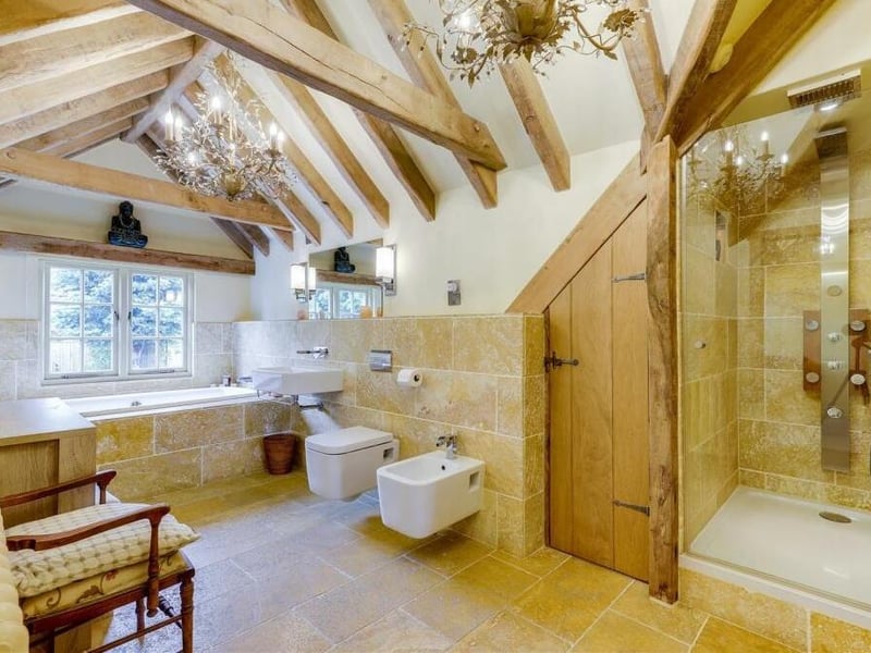 Injecting more luxury to the traditional features of the property is this family bathroom on the first floor. It consists of a bath with tiled surround, a shower enclosure with overhead rainfall shower and massage jets, a wall-mounted wash basin and a low-level flush WC. There is also an in-built storage cupboard, a tiled floor and a half-vaulted ceiling with exposed oak beams and two chandeliers.