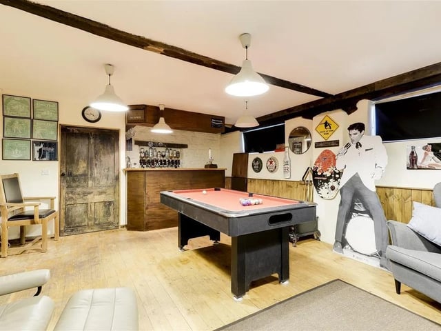 The games room has two windows, a purpose-built bar, lighting and electrics, two wall-mounted heaters, partial wooden paneling on the walls, wood flooring, and a door leading to the garden.