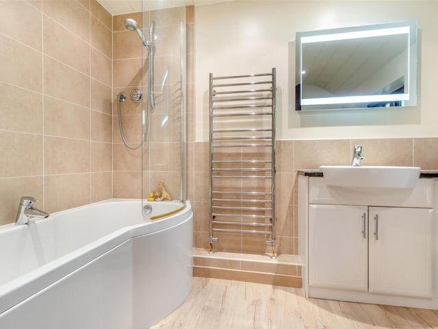 We say farewell to the interior of the Mansfield house with a quick peek inside the family bathroom on the first floor. A three-piece white suite with chrome fittings consists of a P-shaped, panelled bath with mixer tap and shower over, an L-shaped vanity unit with inset wash hand basin, work surfaces to each side and storage cupboards and drawers beneath, and also a low-flush WC. Other features include six ceiling spotlights, a chrome, heated towel-rail and an oak laminate floor.