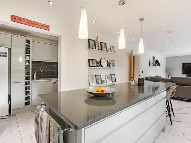 The living area extends seamlessly into this contemporary kitchen, with its large central island or breakfast bar that has a dark grey quartz worktop and high-gloss cabinets and drawers either side. The room boasts a whole host of other cabinets, wall cupboards with under lighting, base units, drawers and worktops.