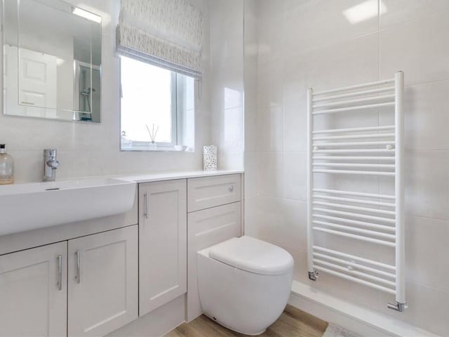 This is the smart en suite bathroom to the second bedroom. A three-piece white suite with chrome fittings features a shower enclosure, vanity unit with inset wash hand basin, and low-flush WC. There are also storage cupboards, a work surface, a heated towel-rail and tiled walls.