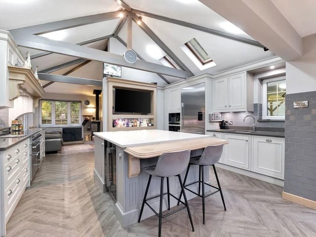 Spruce Lodge is a substantial detached residence extending to approximately 3,714 sq ft in total, set within over two acres of beautifully established woodland and landscaped grounds in one of Ravenshead’s most desirable locations. The kitchen forms the true heart of the home — a striking vaulted space where architectural character meets contemporary functionality. Exposed beams and skylights enhance the sense of volume and flood the room with natural light, while the generous proportions allow it to function as both a practical workspace and a sociable gathering area. At its centre sits a substantial island with quartz worktops and breakfast seating, offering excellent preparation space alongside everyday dining. The shaker-style cabinetry is timeless and beautifully finished, complemented by stone surfaces and quality integrated appliances. An induction range cooker sits beneath a traditional mantle, paired with built-in ovens and an integrated plate warming drawer. A full-height American-style fridge freezer is seamlessly incorporated, while the integrated dishwasher is positioned conveniently beside the sink. Under-cabinet lighting and extensive storage ensure the space is as practical as it is attractive. The kitchen flows naturally into a cosy family snug area, where a contemporary log-burning stove provides warmth and atmosphere.