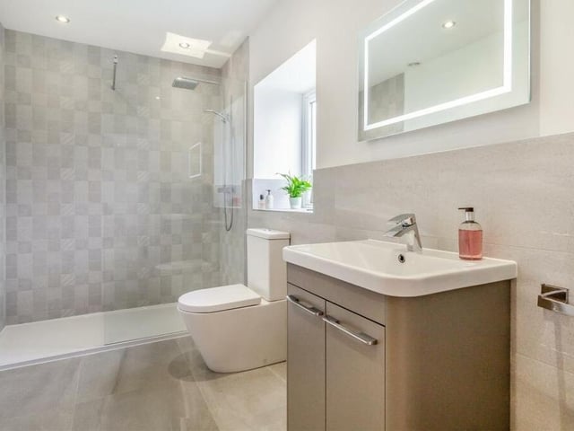 Here is the en suite shower room to the master bedroom. A contemporary three-piece suite with chrome fittings consists of a walk-in, tiled shower enclosure with rainfall shower and additional handset, vanity unit with inset wash hand basin and storage cupboard beneath, and low-flush WC. There is also a fitted mirror, chrome, heated towel rail, modern tiling and ceiling spotlights.