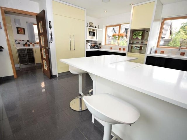 The kitchen includes a practical breakfast bar with seating, polished, tiled flooring and underfloor heating. Bespoke wall and base units offer ample storage, with high-spec Corian worktops and a double sink and drainer. In the background, you can see a utility room off.