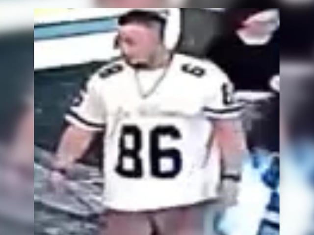 Man with dark hair, goatee, white t-shirt with “86” in black writing, brown shorts.