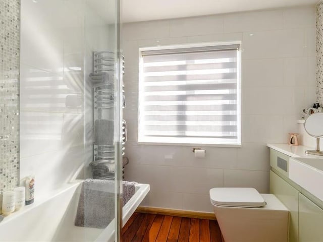 The family bathroom consists of a superbly-appointed, contemporary three-piece white suite with chrome fittings. A P-shaped, panelled bath has a waterfall mixer tap and wall-mounted 'rain' shower above. There is a vanity unit with inset wash hand basin and waterfall mixer tap, plus work surfaces to each side and a storage cupboard beneath. A large fitted mirror above the sink has a mosaic tiling surround.