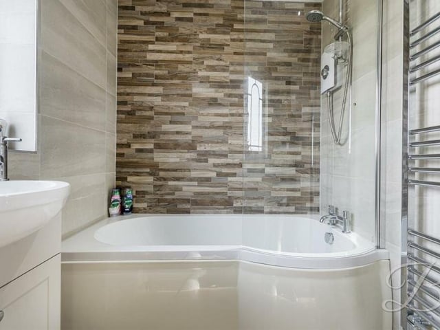 A close-up of the bath, with overhead shower and shower screen, within the family bathroom, which also features a chrome, heated towel-rail.