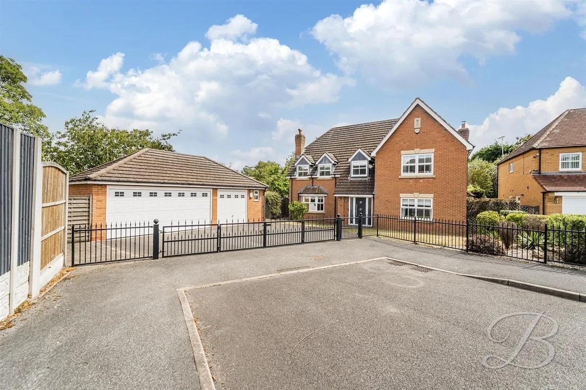 Set on a large plot, the property features a spacious gated entrance leading to a driveway and a large double garage, bordered by a laid lawn and mature shrubbery.