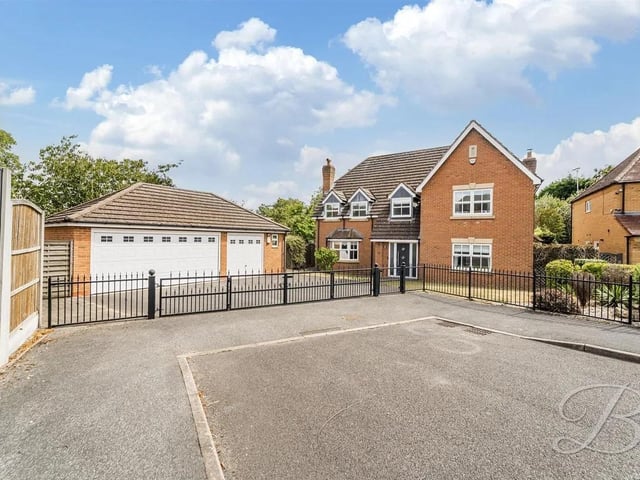 Set on a large plot, the property features a spacious gated entrance leading to a driveway and a large double garage, bordered by a laid lawn and mature shrubbery.