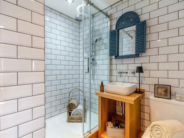 How about this for a stylish en suite shower room to the master bedroom? A three-piece white suite comprises a tiled shower enclosure with rainfall shower and additional handset, wash hand basin, with wall-mounted mixer tap, on an oak table with shelving beneath, and a low-flush WC. There is also a chrome, heated towel-rail, tiled walls and four ceiling spotlights.