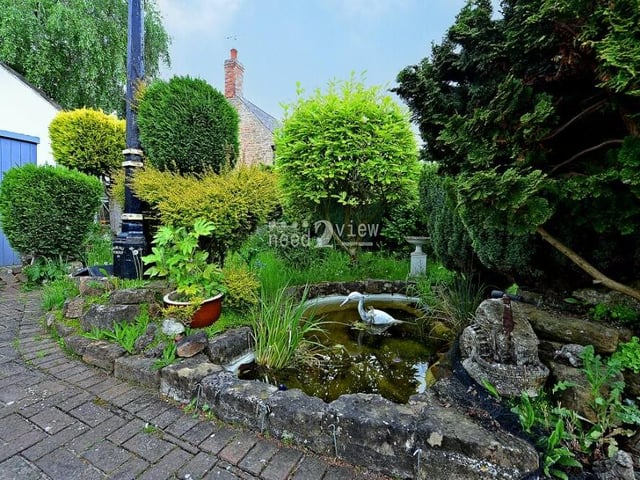 The driveway also leads to this lovely garden pond, which is surrounded by rockery and varied foliage. To the left, there is also an outbulding.