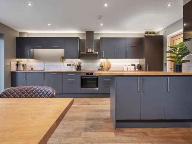 The kitchen features a modern range of subtle dark grey wall cupboards, base units and drawers, with solid wooden work surfaces over. At the heart is an integrated oven and a four-ring induction hob with stainless steel extractor hood over.
