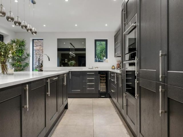 An array of integrated appliances within the kitchen include a Neff slide-and-hide oven, microwave, induction hob with wireless-handled control and inset extractor, wine cooler, full-height fridge and freezer, pull-out larder cupboard and Bosch dishwasher.,