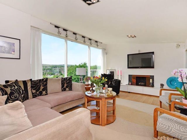 The lounge has a high, sloping ceiling with a floor-to-ceiling picture window. The decor is modern with neutral tones, complemented by a polished oak floor and finished with an inset fire with provision for a TV above and a range of white, gloss fronted units below.