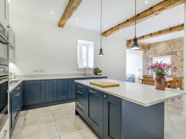 This photo shows the original beams in the L-shaped dining kitchen, which boasts modern shaker cabinets in contrasting colours, wall cupboards, base units and drawers. There is high-quality tiling to the floor and extensive ceiling spotlights.