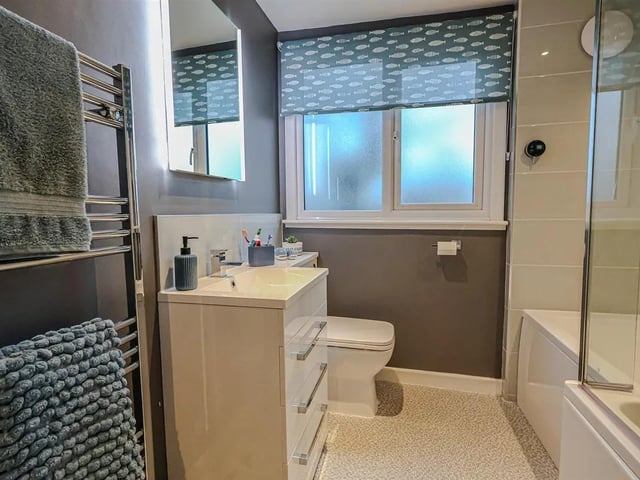 The family bathroom consisting of a shaped bath and screen with full-height tiled surround and mains shower with external pre-heat control, wash basin and concealed cistern toilet set into a vanity surround with drawers.
