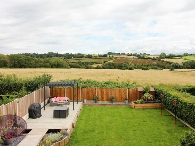 This image shows not only the top end of the back garden,with its paved patio, plants and shrubs, but also the open, rolling countryside that stretches into the distance.