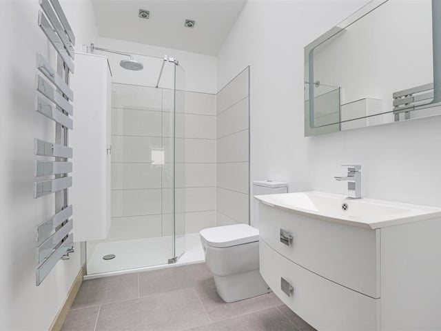 The en suite room to the second bedroom is, as you can see, as clean as a whistle. It is dominated by a shower enclosure with overhead rainfall shower, but there is also a wall-mounted storage unit, wash basin with storage drawers, low-level, dual-flush WC, a chrome, heated towel-rail, recessed spotlights, underfloor heating and a skylight window.