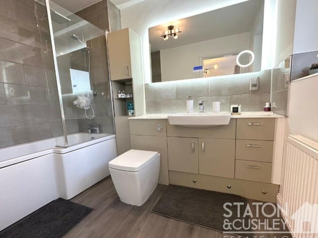 This is the en suite bathroom, accessible from the master bedroom on the ground floor. It includes a P-shaped, panelled bath with overhead mains shower, mixer tap and shower screen, as well as a vanity unit with mounted sink, low-level WC and wall-mounted mirror, plus storage.