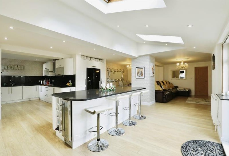 The first room to look at is the modern, fitted high-gloss kitchen in one of the properties. It features a range of units, black granite work surfaces, breakfast bar with granite top, an integrated double oven, microwave and steam oven, plus an induction hob with hood and extractor over. A pillar separates a dining and living area.
