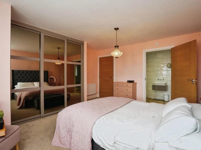 The master bedroom is spacious and peaceful, decorated in soft pink tones with carpeted flooring. An en-suite bathroom is accessed via a wooden door, adding convenience and privacy.