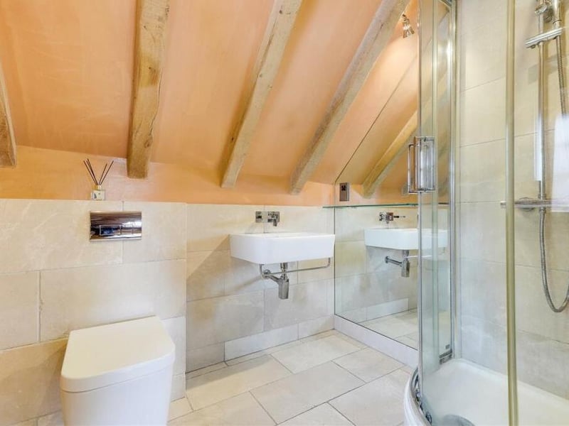 One of the first-floor bedrooms has access to this excellent en suite shower room. It includes a shower enclosure with overhead rainfall shower, wall-mounted wash basin, low-level flush WC, heated towel-rail, exposed oak beams to the ceiling, partly tiled walls and a tiled floor.
