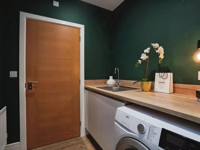 The utility room continues the kitchen's dark green walls and wood-effect flooring. It houses a washing machine beneath a wood-effect work surface with a sink and offers additional cupboard space. A door leads into the kitchen/dining area, making household chores more convenient and well organised